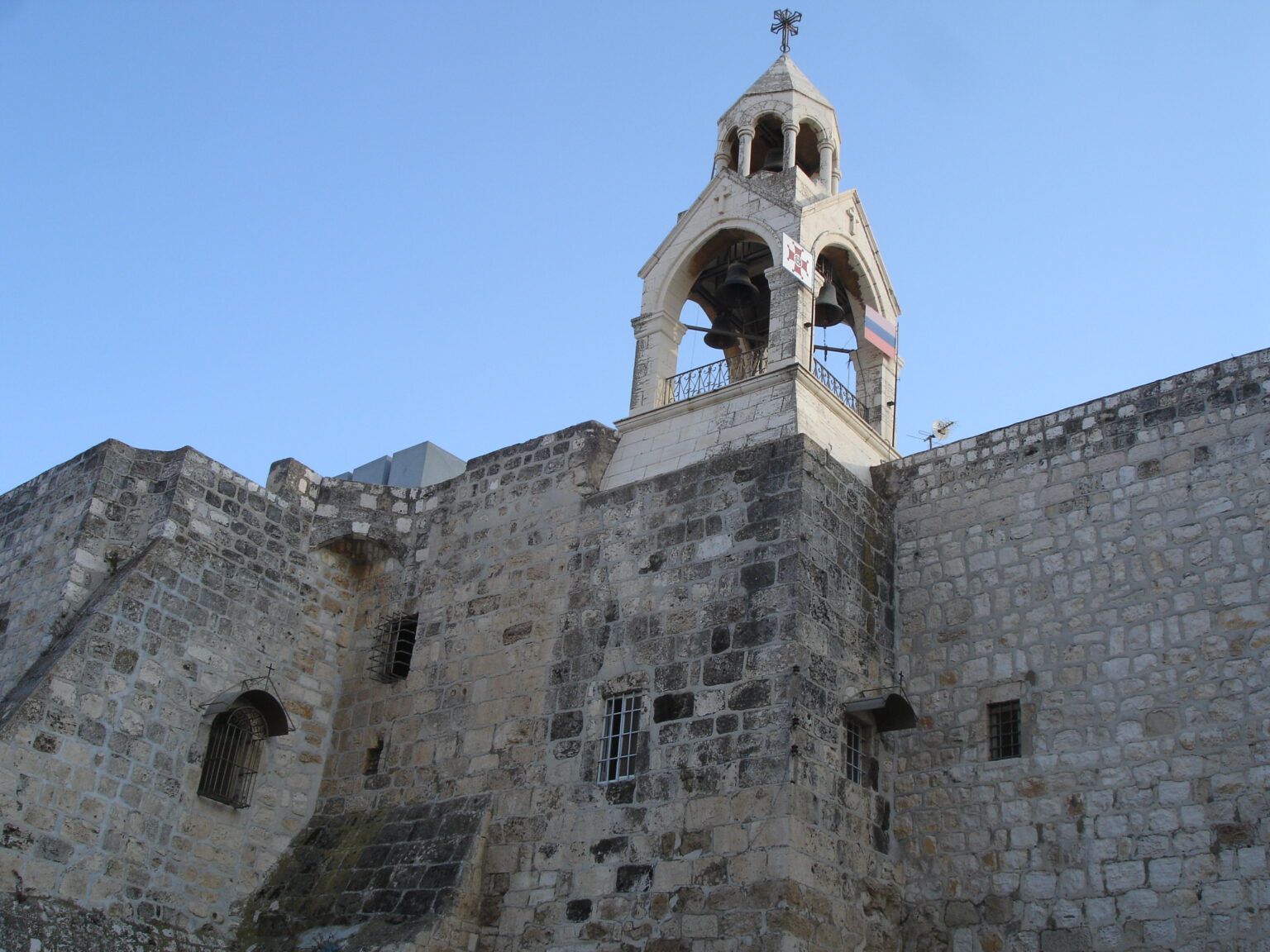 Campanário de pedra da Igreja da Natividade, com sinos e cruz no topo, acima de paredes de pedra sob céu azul, em Belém.