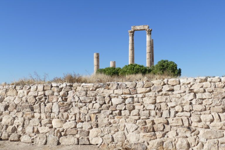 Colunas de pedra do Templo de Hércules, na Cidadela de Amã, acima de uma muralha e arbustos sob céu azul.
