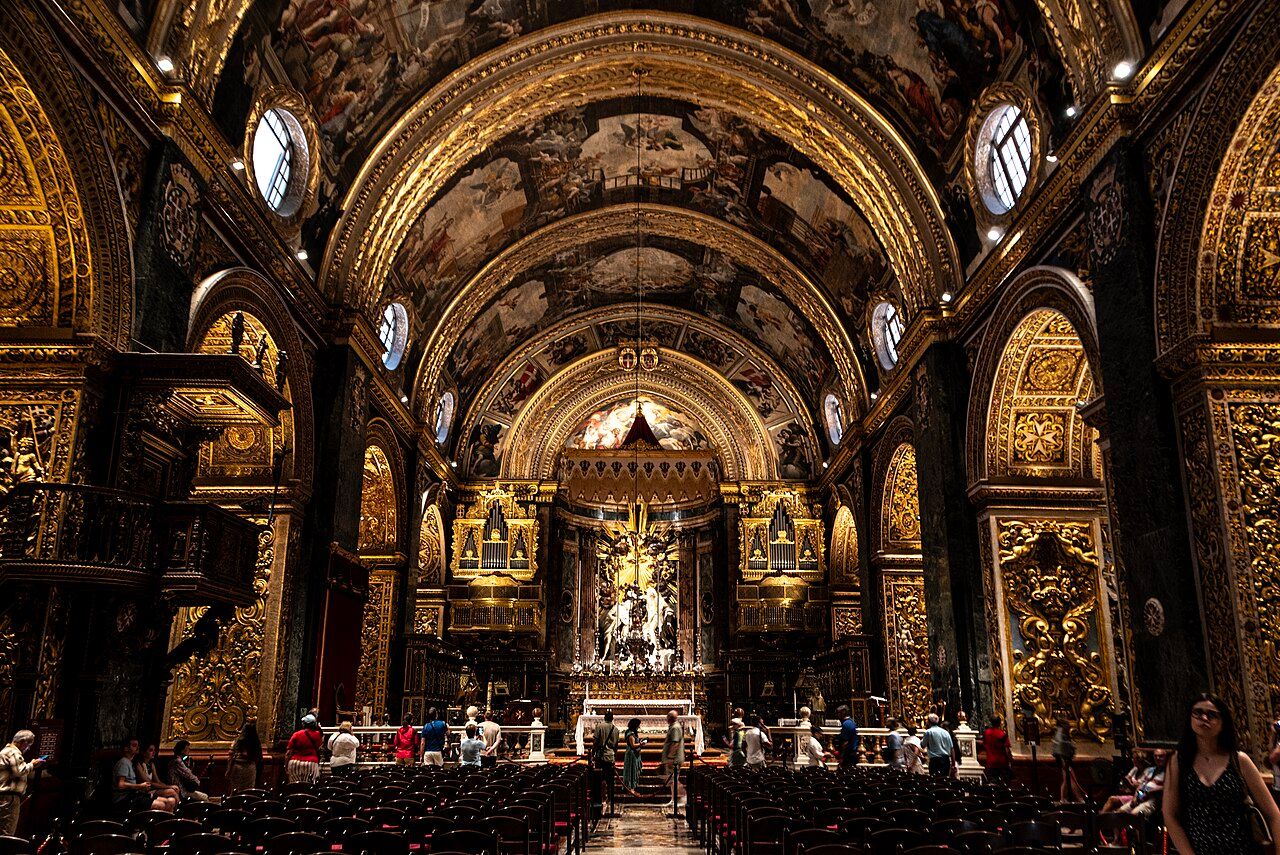Vista da nave da Co-Catedral de São João, com capelas laterais, abóbada pintada e visitantes.