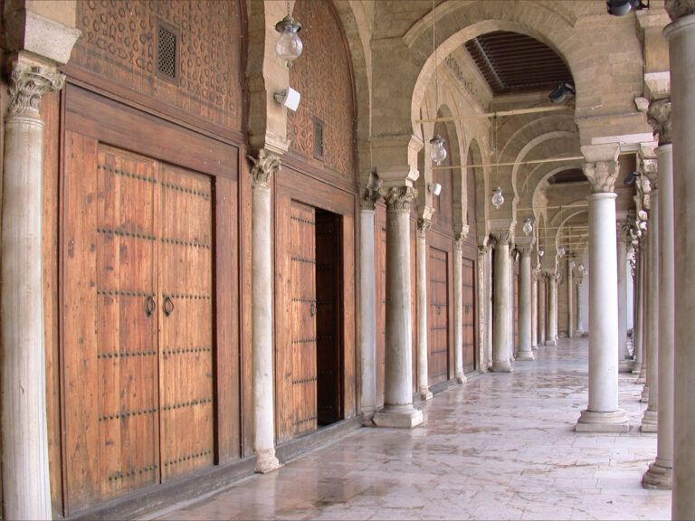 Galeria da Mesquita Zitouna, com grandes portas de madeira à esquerda e uma sequência de colunas claras e arcos à direita.
