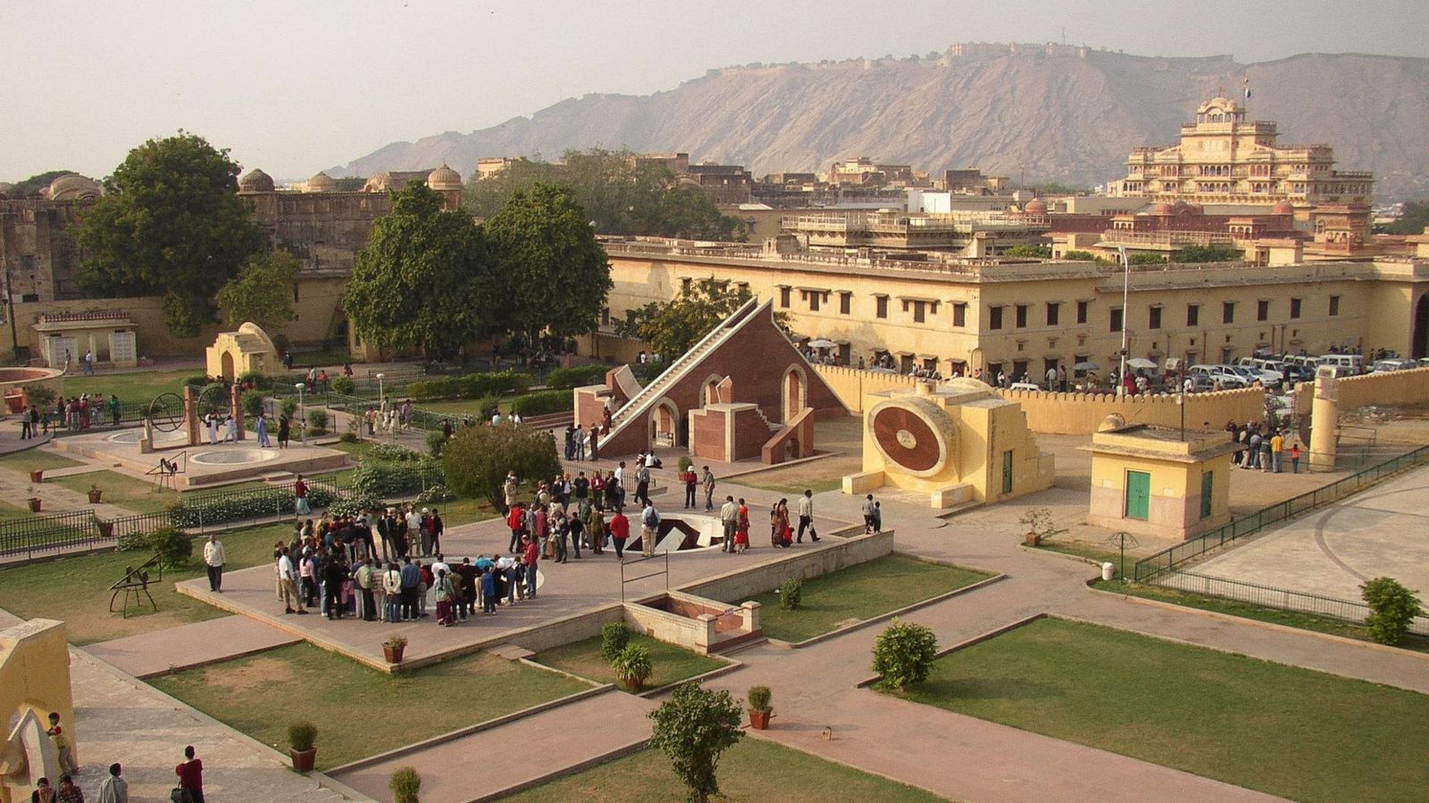 Vista elevada dos instrumentos e jardins do Jantar Mantar em Jaipur, com visitantes e edifícios ao fundo, em 2002.