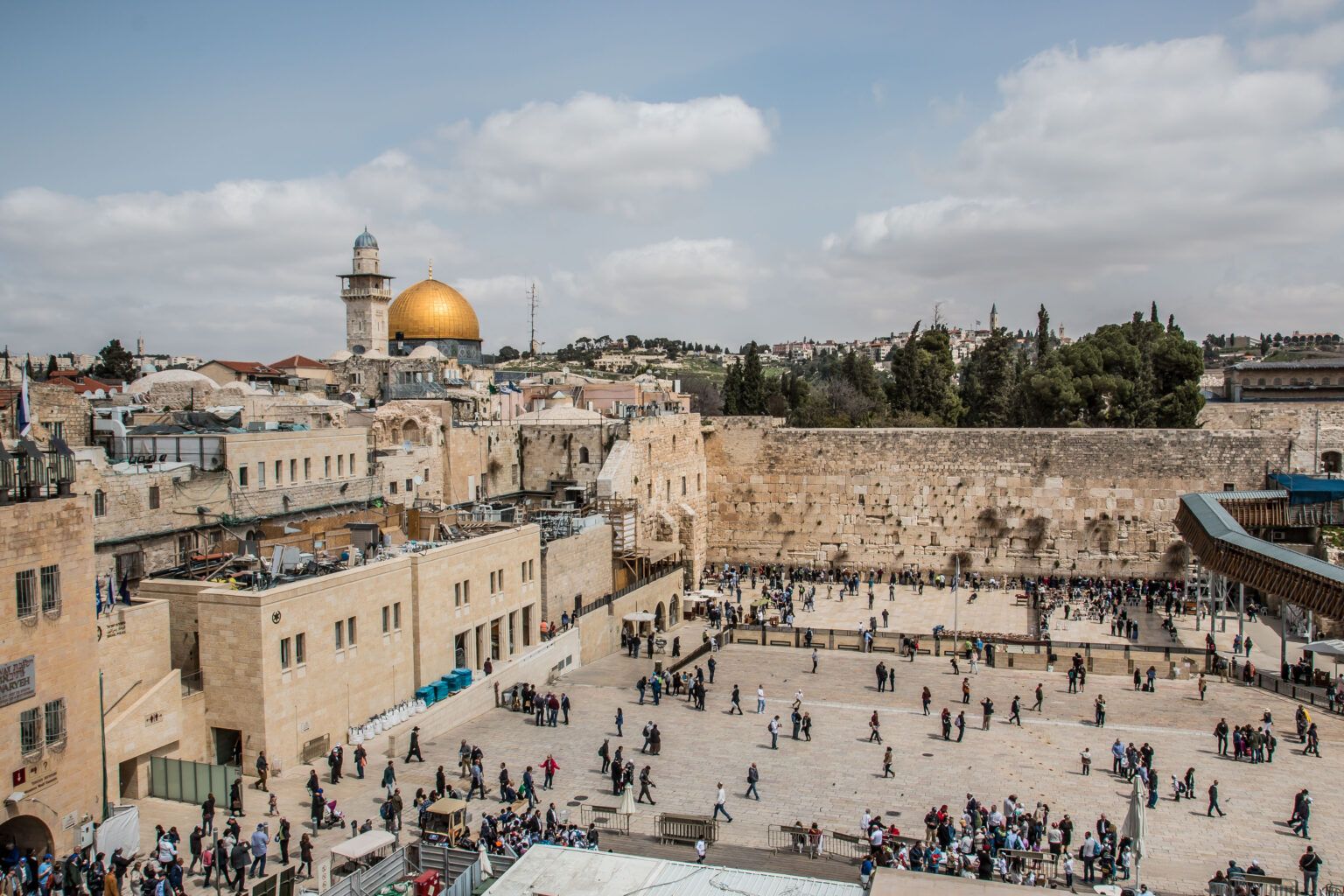 Vista da praça do Muro Ocidental com visitantes, edificações de pedra e a cúpula dourada do Domo da Rocha ao fundo, em Jerusalém Oriental.
