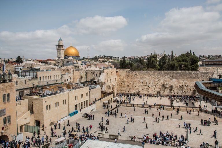Vista da praça do Muro Ocidental com visitantes, edificações de pedra e a cúpula dourada do Domo da Rocha ao fundo, em Jerusalém Oriental.