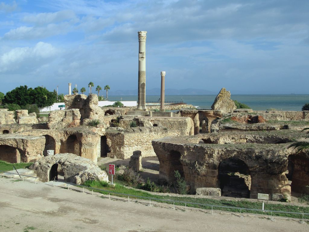 Ruínas das Termas de Antonino com passagens arqueadas, duas colunas altas ao centro e o mar ao fundo, em Cartago.