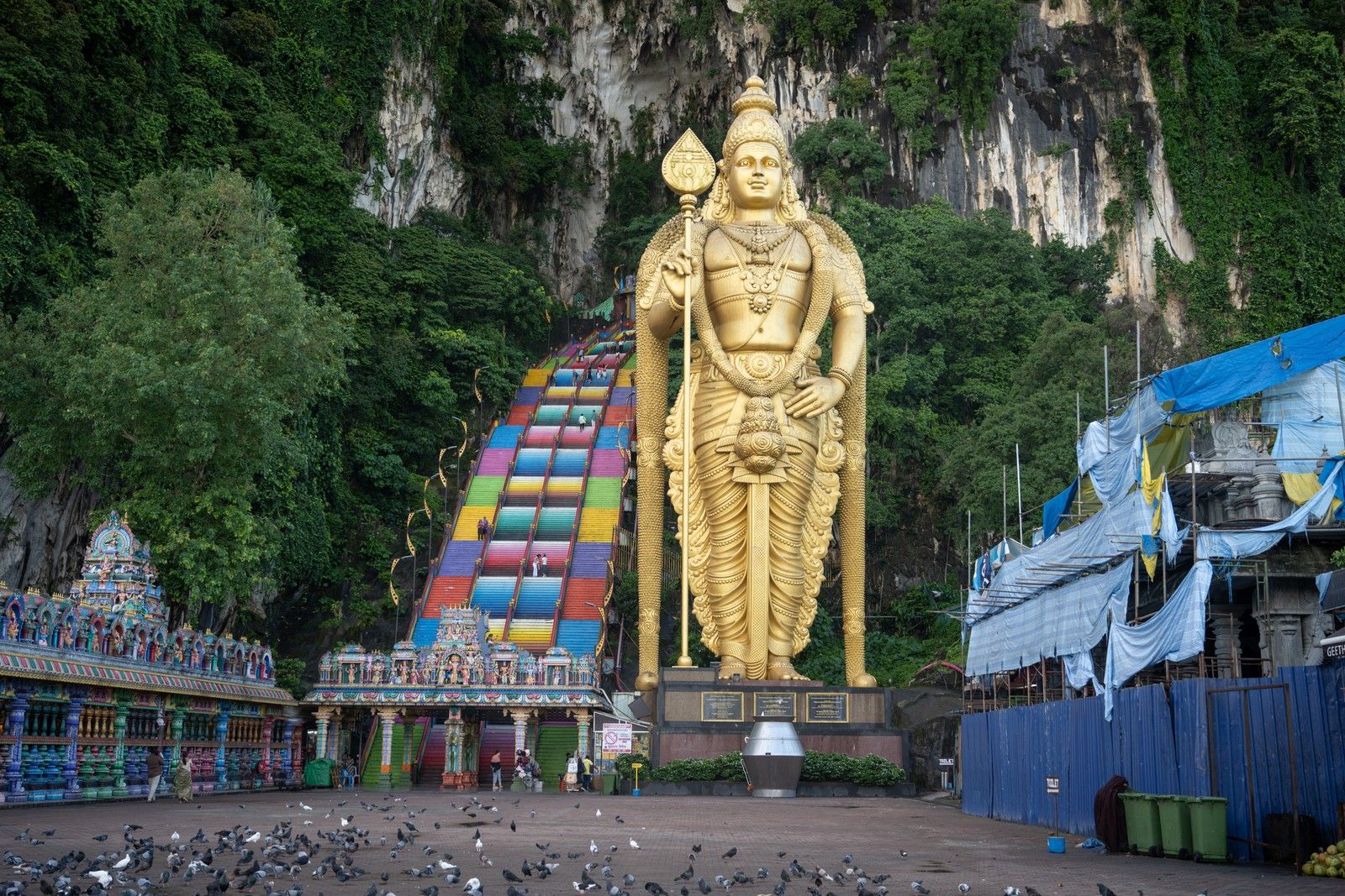 Estátua dourada de Murugan diante das falésias de Batu Caves, com escadaria colorida à esquerda, pombos na praça e estruturas cobertas por lonas à direita.