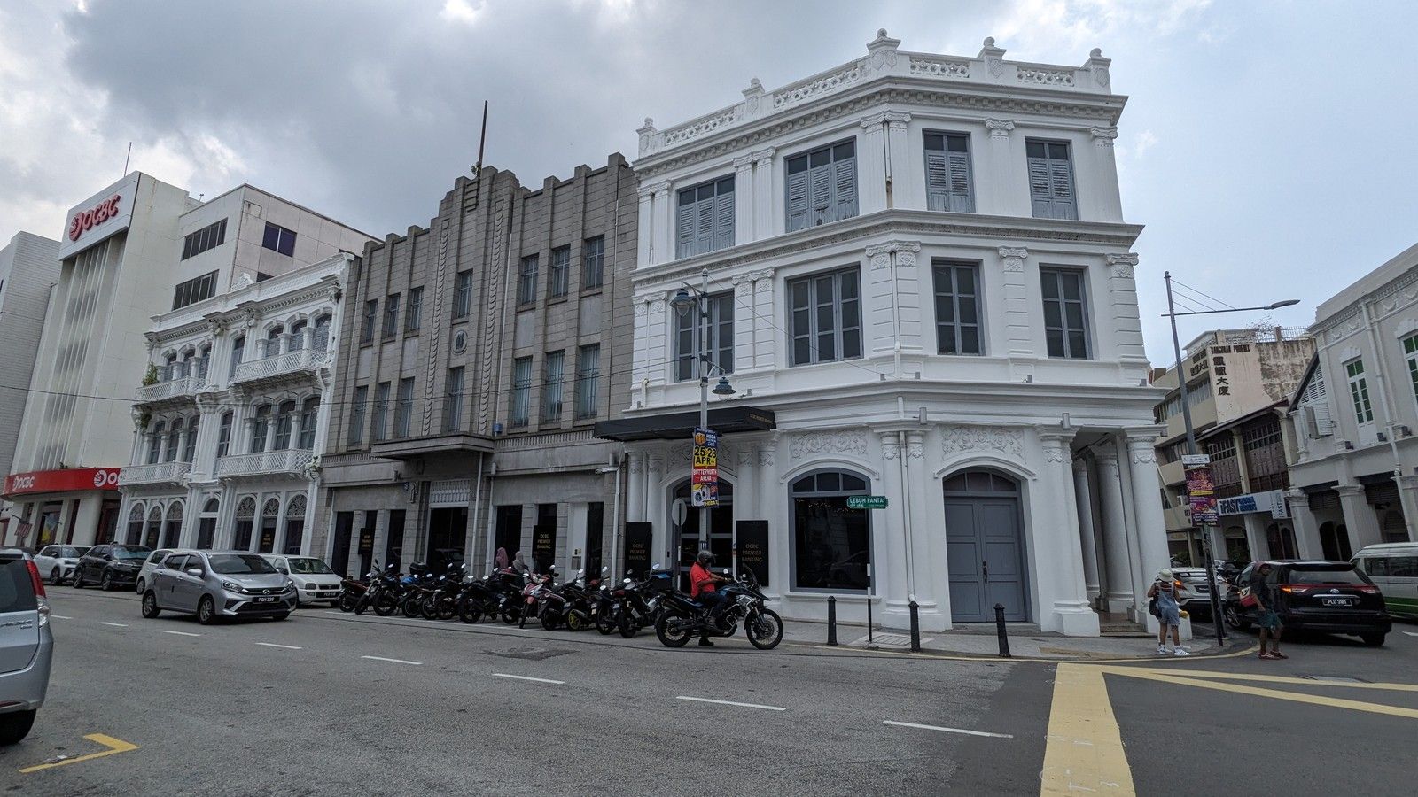 Fachadas históricas brancas e cinzentas na Lebuh Pantai, em George Town, com veículos e pedestres junto à rua, sob céu nublado.
